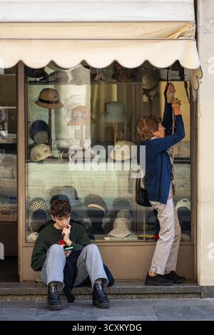 Un giovane uomo si siede premurosamente vicino a un negozio di cappelli, mentre una donna più anziana adatta la tenda in una strada tranquilla. Foto Stock