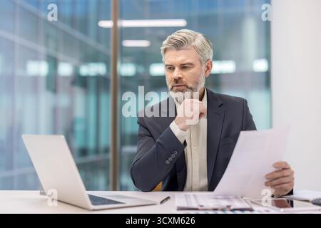 Uomo d'affari maturo con la barba che esamina attentamente documenti e dati finanziari in un ufficio aziendale, utilizzando un laptop, pensando a importan Foto Stock