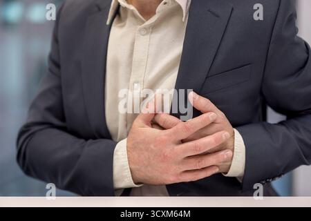 Uomo in giacca e camicia che stringe il petto con entrambe le mani, avverte dolore acuto al petto, un sintomo di potenziali problemi cardiaci o un cuore a. Foto Stock