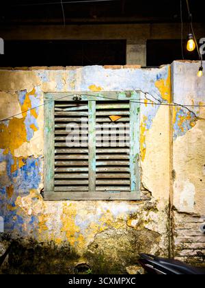 Vecchie pareti intemprate con vernice sbucciante e una rustica finestra con persiane in legno, illuminata da lampadine appese Ipoh malesia - persiane Foto Stock