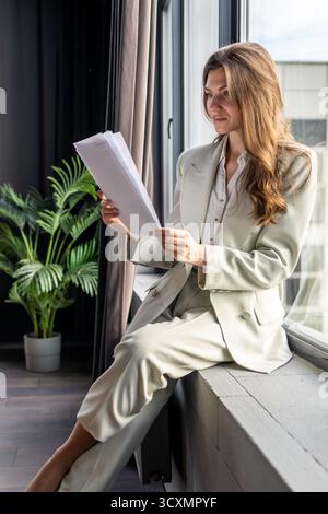 Donna d'affari che esamina contratti importanti mentre siede su un davanzale nel suo ufficio moderno, concentrandosi su dettagli importanti per il successo finanziario A. Foto Stock
