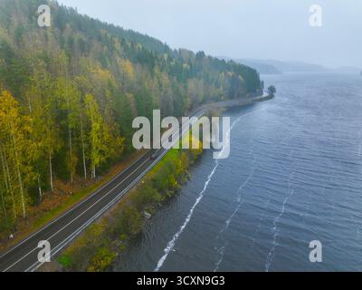 Vista aerea di una piccola auto rossa che viaggia lungo una tortuosa strada asfaltata delimitata da un ampio lago e da una foresta mista dai vivaci colori autunnali Foto Stock