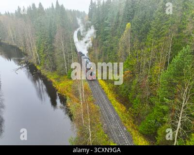 Vista aerea di una locomotiva a vapore nera e rossa con fumo che ondeggia, viaggiando su una pista accanto a un fiume in una foresta nebbiosa Foto Stock