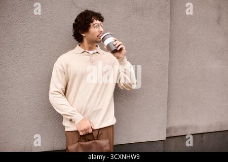 Un giovane sorseggia il caffè con cura, vestito in modo informale con tonalità calde, contro un muro elegante. Foto Stock
