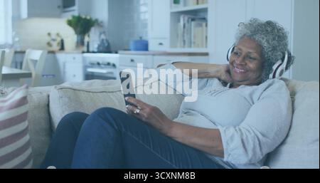 Donna anziana sdraiata che indossa le cuffie tenendo lo smartphone sul divano vicino alla cucina aperta, spazio per le copie Foto Stock