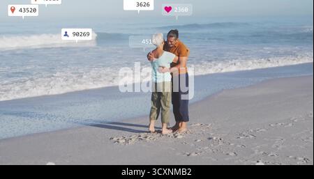 Abbracciando una coppia senior in piedi su una spiaggia sabbiosa in riva al mare, con sovrapposizioni digitali galleggianti Foto Stock
