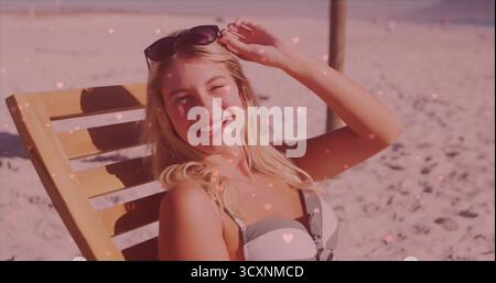 Donna che indossa un paio di occhiali da sole regolabili sulla sedia da spiaggia in riva al mare con cuori rosa, spazio fotocopie Foto Stock