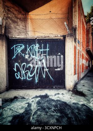 Vecchia porta del garage con graffiti blu in un vicolo urbano circondato da pareti in mattoni e ombre dure Foto Stock
