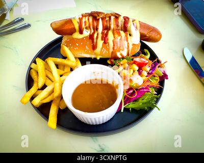 Hot dog gourmet con senape e ketchup, servito insieme a patatine fritte croccanti, insalata fresca e ricca salsa da immersione su piatto nero nel Cafe malaysia Foto Stock