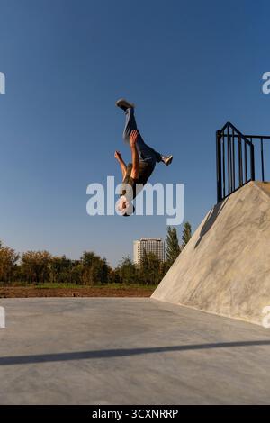 Il freerunner è stato catturato capovolto a mezz'aria mentre si capovolge da un pendio di cemento, congelato in posizione verticale contro il cielo limpido in un intenso movimento urbano Foto Stock