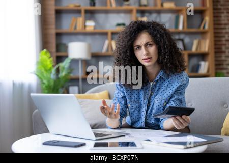 Donna confusa seduta su un divano che calcola le spese mensili con una calcolatrice e un notebook, che lotta con le finanze e sembra stressata durante la gestione Foto Stock