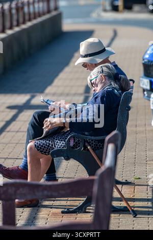 una coppia più anziana si gode un pomeriggio rilassante seduto su una panchina al mare in una giornata di sole, una signora anziana che si addormenta sulla spalla del marito al sole. Foto Stock