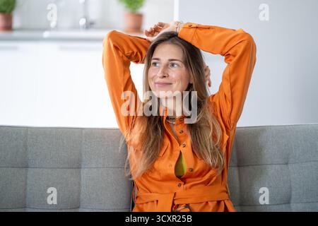 Donna di mezza età in stato onirico che guarda da parte trasmettendo una profonda contemplazione seduta sul divano di casa Foto Stock