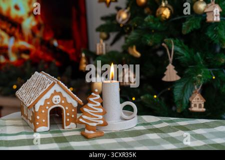 Casa del pan di zenzero e biscotti all'albero di Natale con glassa bianca accanto alla candela in fiamme sul tavolo festivo con albero decorato. Concetto di cottura per le vacanze t Foto Stock