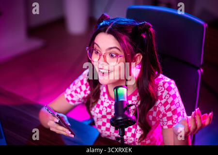 Adorabile blogger donna che ospita un coinvolgente live streaming sui social media con illuminazione vivace e microfono in uno spazio di lavoro creativo Foto Stock