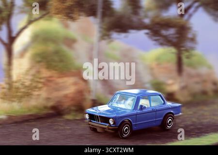 l'auto blu in stile vintage viaggia lungo una strada strutturata con uno sfondo sfocato di alberi e grandi rocce. La composizione evoca un viaggio nostalgico, b Foto Stock