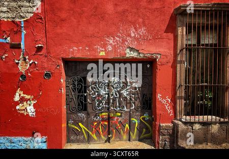 Una parete rossa intemprata con vivaci graffiti che coprono un cancello metallico, che mostra arte urbana in un ambiente rustico di quartiere Foto Stock