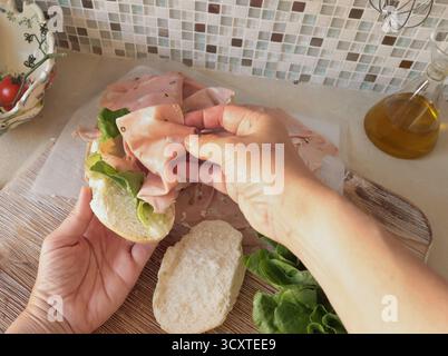 Una persona anonima prepara un panino con salsiccia, lattuga e mortadella, maionese fatta in casa in cucina. Cucina italiana, alta qualità coltivata in azienda Foto Stock