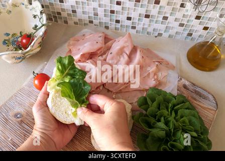 Una persona anonima prepara un panino con salsiccia, lattuga e mortadella, maionese fatta in casa in cucina. Cucina italiana, alta qualità coltivata in azienda Foto Stock