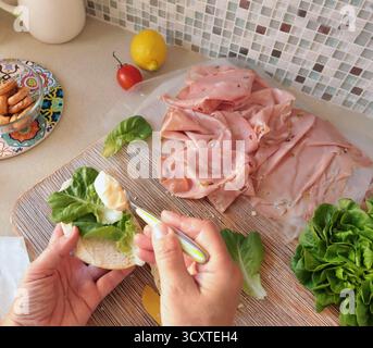 Una persona anonima prepara un panino con salsiccia, lattuga e mortadella, maionese fatta in casa in cucina. Cucina italiana, alta qualità coltivata in azienda Foto Stock