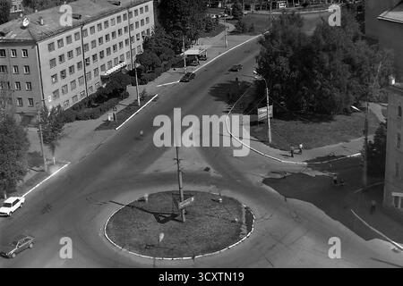 Una foto d'archivio ad alto angolo degli anni '1980 cattura un incrocio affollato a Sloviansk, RSS Ucraina. La vista si concentra su una rotatoria in via Yunykh Kommunarov (ora via Bankivska), con l'edificio del Slavyansk Aviation Technical College sulla sinistra. Le classiche auto dell'epoca sovietica e un ciclista navigano all'incrocio, mentre i pedoni camminano lungo i marciapiedi. Questa istantanea del traffico urbano quotidiano è un prezioso resoconto della vita di routine in un tranquillo Donbas prima della guerra Foto Stock