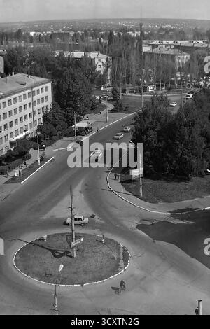 Una foto d'archivio ad alto angolo degli anni '1980 cattura un incrocio affollato a Sloviansk, RSS Ucraina. La vista si concentra su una rotatoria in via Yunykh Kommunarov (ora via Bankivska), con l'edificio del Slavyansk Aviation Technical College sulla sinistra. Le classiche auto dell'epoca sovietica e un ciclista navigano all'incrocio, mentre i pedoni camminano lungo i marciapiedi. Questa istantanea del traffico urbano quotidiano è un prezioso resoconto della vita di routine in un tranquillo Donbas prima della guerra Foto Stock
