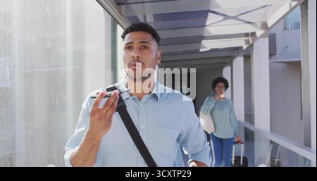 Uomo ispanico che parla allo smartphone in skybridge, con donna che beve caffè e tira la valigia Foto Stock
