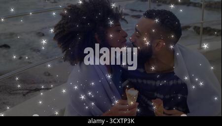 Coppia pendente che tiene in mano coni di vaniglia che si fondono sulla sporgenza dal mare, avvolta in coperta vicino a ringhiere metalliche Foto Stock