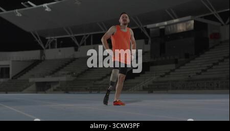 Atleta in piedi che indossa la parte superiore arancione, utilizzando la lama protesica sulla pista blu sotto i riflettori Foto Stock