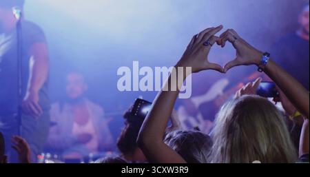 Donna che si forma a cuore e indossa un top senza maniche in un locale di musica dal vivo, con telefoni e luci da palcoscenico Foto Stock