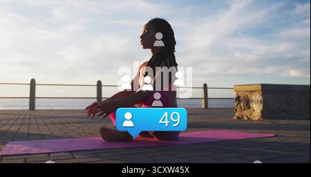 Donna seduta che indossa abbigliamento sportivo rosa che meditava sul tappetino yoga rosa sul lungomare, con ringhiera Foto Stock