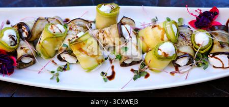preparare involtini vegetariani di zucchine e melanzane con formaggio feta e ripieno di semi di zucca arrostiti disposti su un piatto bianco con verdure fresche e colorate. moderno ambiente da cucina del ristorante. Foto Stock