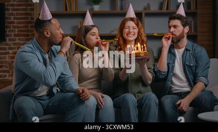 Quattro amici multirazziali che cantano una canzone di buon compleanno alla donna caucasica che tiene una torta festosa con candele sorridenti e soddisfatti congratularsi con la ragazza a casa Foto Stock