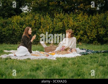 Due donne sono sedute su una coperta in un parco, mangiano cibo e si godono la compagnia l'una dell'altra Foto Stock