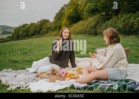 Due donne sono sedute su una coperta in un campo erboso, mangiano cibo e si godono la compagnia reciproca Foto Stock