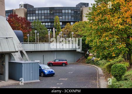 Rampa di parcheggio curva con scarabeo Volkswagen blu e SUV rosso parcheggiati sotto il ponte Gray Overpass, circondato da vibranti alberi autunnali e moderni edifici Foto Stock