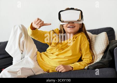 Una ragazza alfa-gen si rilassa sul divano di casa, indossando un visore VR e sorridendo felicemente. Foto Stock