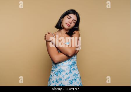 Una donna sta in piedi con fiducia a occhi chiusi, abbracciandosi con un abito floreale blu. Lo sfondo neutro esalta la sua espressione di auto-amore e. Foto Stock