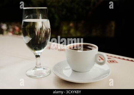 Tazza in porcellana bianca con forte caffè turco nero o americano e un bicchiere d'acqua servito su un tavolo da caffè all'aperto con luce naturale. Foto Stock