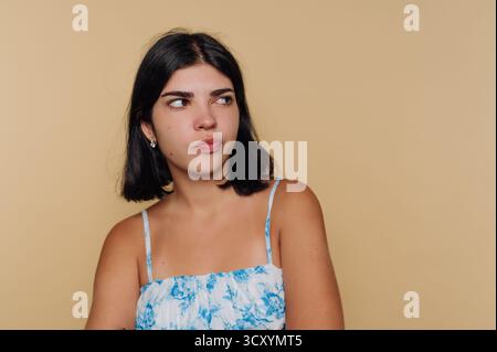 Una giovane donna con i capelli scuri a spalla indossa un top floreale e si siede davanti a uno sfondo liscio e abbronzato. Guarda con attenzione, riflettendo una moma Foto Stock