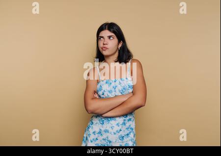 Una giovane donna sta in piedi con le braccia incrociate, mostrando un aspetto scettico. Indossa un abito floreale ed è in una stanza semplice con uno sfondo di colore neutro Foto Stock