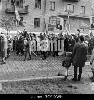 Una suggestiva foto d'archivio del 7 novembre 1974 cattura lo spirito di una parata dell'anniversario della rivoluzione d'ottobre a Sloviansk, RSS Ucraina. Una grande colonna di manifestanti, comprese famiglie con bambini, marcia lungo una strada acciottolata. I partecipanti portano bandiere sovietiche, ritratti di leader politici, striscioni con slogan come "Glory to the Heroes of Labor" e palloncini festivi. Questa immagine è un vivido documento di una grande festa di stato e della vita pubblica di un pacifico Donbas prima della guerra Foto Stock