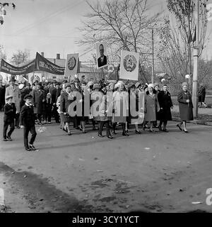 Una suggestiva foto d'archivio del 7 novembre 1974 cattura una dimostrazione su larga scala per l'anniversario della Rivoluzione d'ottobre a Sloviansk. Colonne di cittadini, comprese famiglie e una banda di ottoni, marciano attraverso le strade e la piazza centrali della città. Portano con orgoglio bandiere sovietiche, striscioni e grandi ritratti di Lenin e di altri leader politici. Questa immagine è un vivido documento di una grande festa di stato, che mostra lo spirito collettivo e la vita pubblica di un pacifico Donba prima della guerra Foto Stock