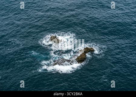 Prospettiva aerea minimalista di formazioni rocciose isolate circondate da schiuma bianca nelle acque blu profonde dell'oceano, sfondo naturale ideale e astratto Foto Stock