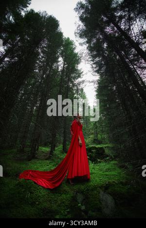 la donna che indossa un lungo abito rosso si erge graziosamente in una lussureggiante foresta verde. alberi alti la circondano, creando un'atmosfera mistica durante il crepuscolo. cen Foto Stock