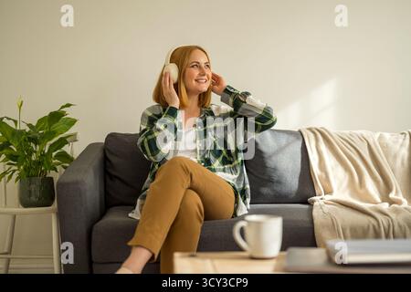 Donna che ascolta musica con le cuffie sedute su un divano Foto Stock