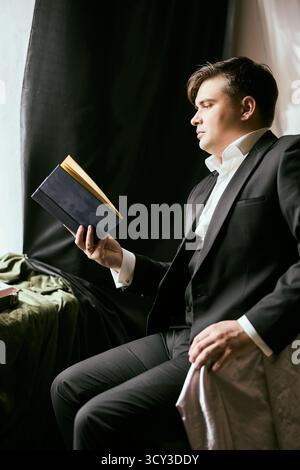 Uomo in abito formale che legge un libro vicino alla finestra in un ambiente caldo Foto Stock