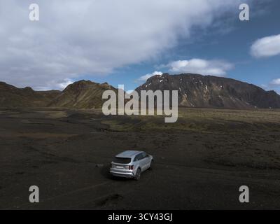 Veduta aerea di un SUV solitario parcheggiato sulla terra vulcanica nera, sullo sfondo di aspre montagne sotto un vasto cielo, Reykjavík, Islanda. Foto Stock