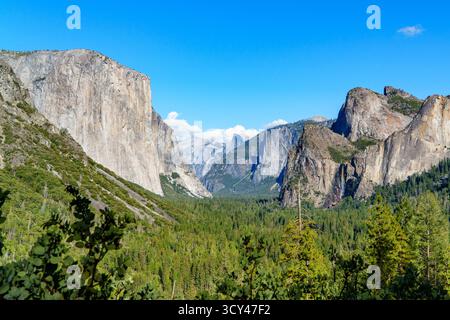 Un'ampia vista della valle di Yosemite mostra iconiche formazioni di granito come El Capitan, Half Dome e lussureggianti foreste verdi sotto un cielo azzurro. Foto Stock