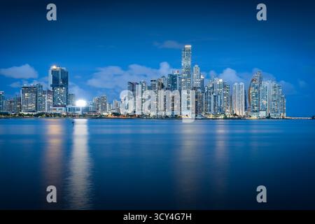Vista panoramica del moderno skyline di Panama City all'ora blu, con grattacieli illuminati che si riflettono sul mare calmo, che mostrano la vibrante urba Foto Stock